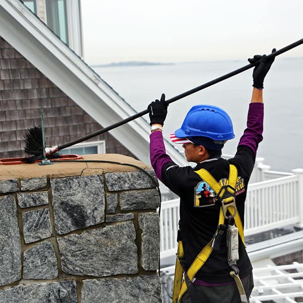 A professional chimney sweep in Framingham, MA wearing safety gear uses rotary brushes and vacuum equipment to clean soot and creosote from a residential chimney.