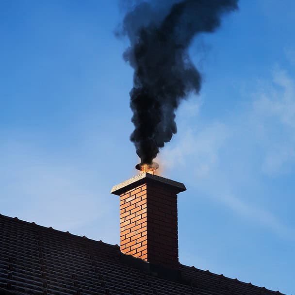 A brick chimney on a roof in Norfolk County, MA, emits a thick column of black smoke against a clear blue sky, highlighting the need for expert chimney repairs.