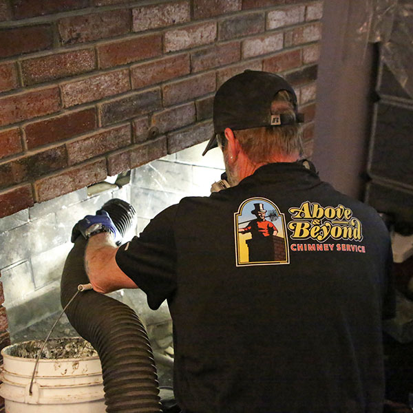 A chimney technician wearing an "Above & Beyond Chimney Service" shirt uses a vacuum hose to clean a brick fireplace in Massachusetts, collecting soot and debris in a white bucket—delivering trusted chimney sweep services across Norfolk and Plymouth County.
