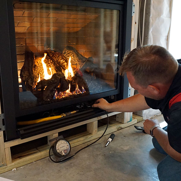 A person is adjusting a gas fireplace with logs inside, showing tall flames. Nearby, a pressure gauge and tools hint at recent chimney services in Norfolk & Plymouth County. The fireplace is set in a partially constructed wall.