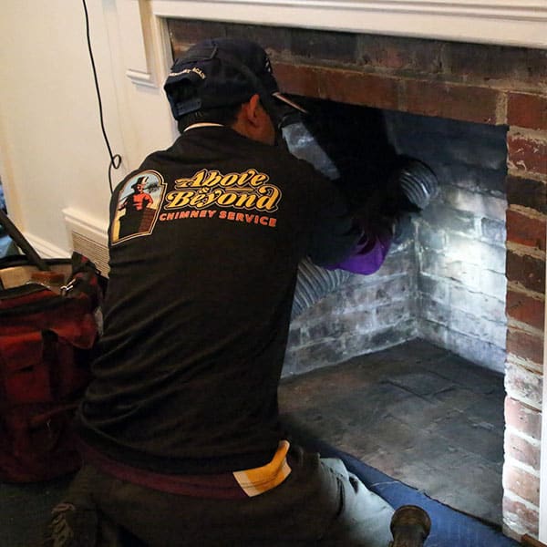 A person donning an "Above & Beyond Chimney Service" shirt cleans the inside of a brick fireplace, kneeling with tools scattered nearby. Only their back is visible as they work diligently, showcasing expertise in chimney repairs across Norfolk & Plymouth County, MA. White walls and a small light illuminate the space.