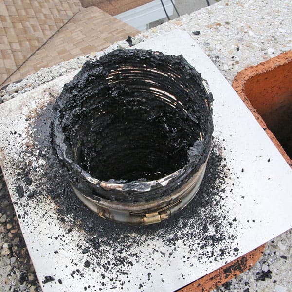 A chimney flue viewed from above, heavily coated with black soot and creosote buildup, signals the need for cleaning or chimney repairs in Norfolk & Plymouth County, MA. The circular metal pipe shows extensive residue clinging to its inner surface, while surrounding bricks are partially visible.