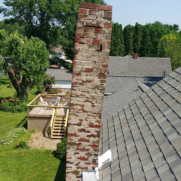 A brick chimney leans precariously on the shingled rooftop, awaiting essential chimney repairs in Norfolk & Plymouth County. The backdrop reveals a deck, lawn, and trees under the clear MA sky.