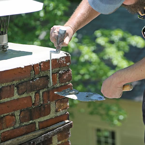 A person expertly repairing a brick chimney with a trowel and level. Sunlight illuminates the bricks and fresh mortar, creating a warm glow. Green foliage provides a serene backdrop, emblematic of the idyllic settings found in Norfolk & Plymouth County, MA.