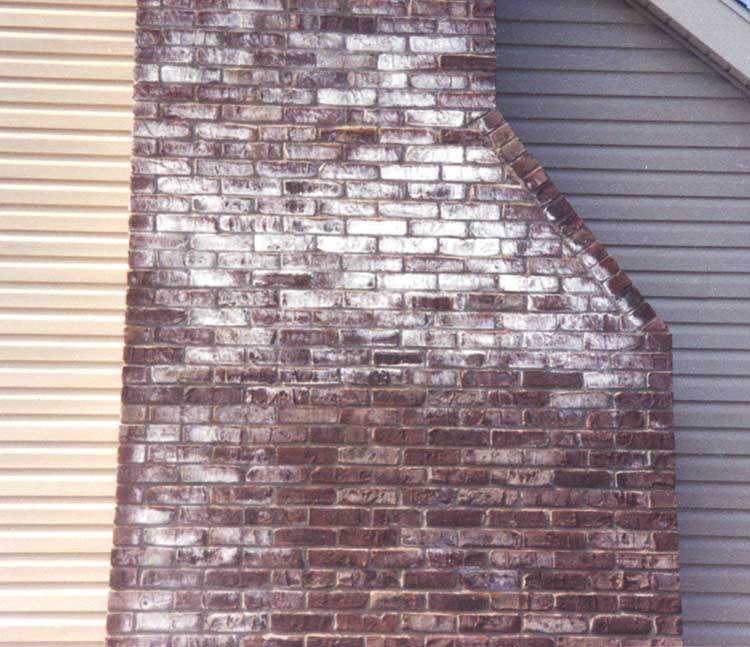 A weathered brick chimney contrasts with adjacent horizontal siding in Norfolk & Plymouth County, MA. Discoloration from exposure is evident on the bricks, while the siding on each side is beige and gray. The chimney's stepped pattern highlights the need for expert chimney services.