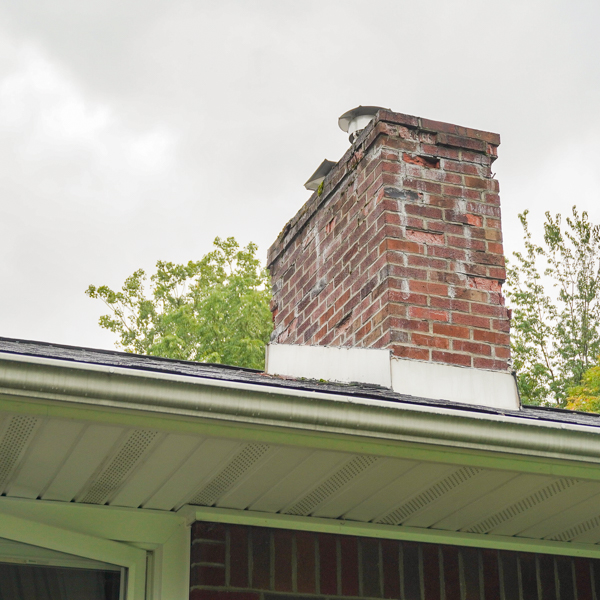 A damaged red brick chimney, with visible bricks missing and cracks near the top, stands above a house roof in MA. The gray roof features a white gutter, set against trees under an overcast sky. Consider chimney services Norfolk & Plymouth County for restoration expertise.