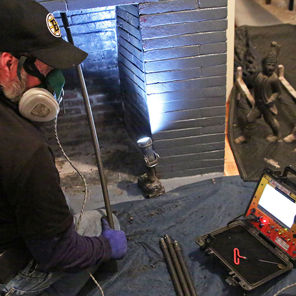 A technician, donned in a mask and gloves, carefully examines a chimney for the renowned Chimney Services Norfolk & Plymouth County. Using a flashlight and camera, the internal view is displayed on a monitor. Nearby, a statue of a chimney sweep stands as a nod to tradition.