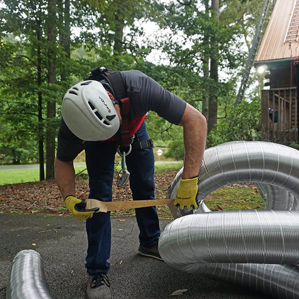 A person wearing a safety helmet and gloves bends over, measuring a long, flexible metal duct on the ground. They are surrounded by trees and grass, with a wooden structure visible in the background—an essential task in chimney services across Norfolk & Plymouth County.