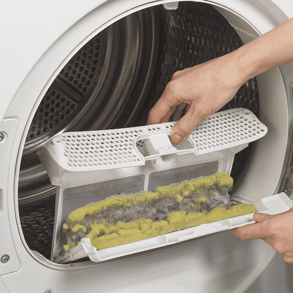 A person removes a lint filter from a dryer, revealing accumulated lint much like the residue found in chimneys needing cleaning. The drum of the dryer is visible in the background, reminiscent of reliable chimney services in Norfolk & Plymouth County, with lint a mix of gray and yellow fibers.