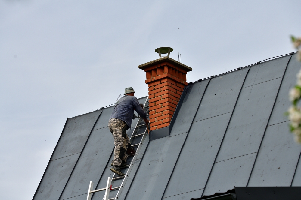 A person in camouflage pants and a hat climbs a ladder leaned against a steep metal roof, working near a red brick chimney under a clear MA sky—possibly performing chimney restoration in Norfolk and Plymouth County.
