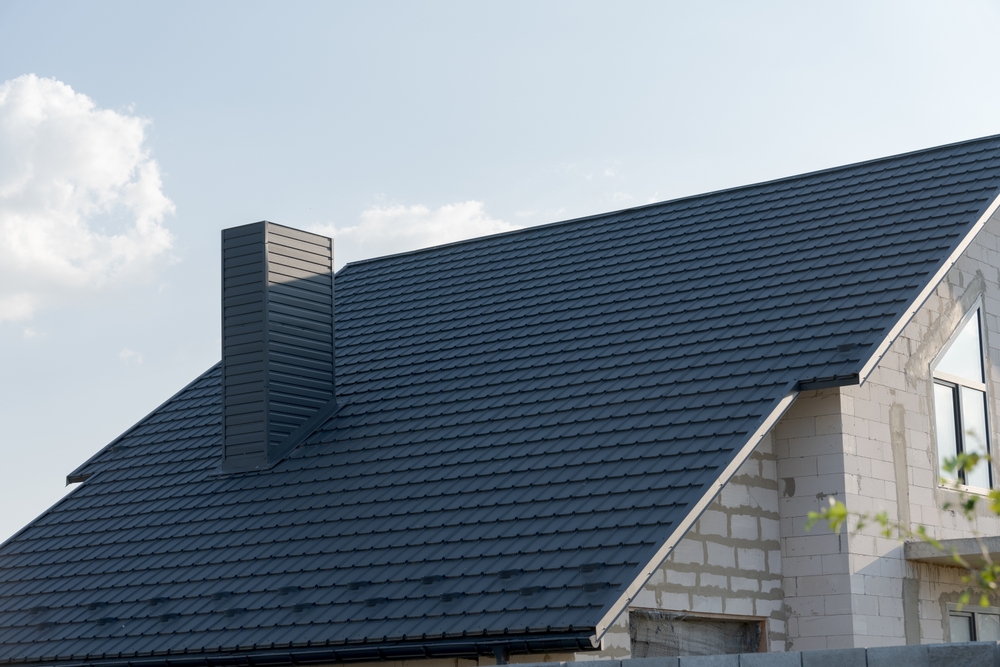 A modern house in MA with light-colored brick walls and a dark gray metal shingle roof, featuring a prominent rectangular chimney—ideal for those seeking chimney repair in Norfolk and Plymouth County, MA—stands under a clear sky.