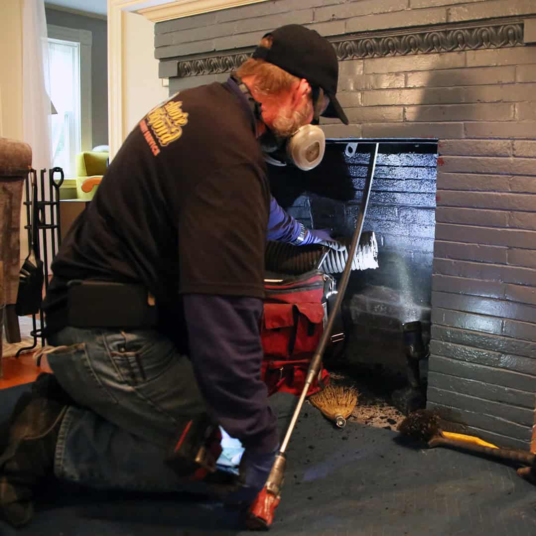 A professional chimney technician in uniform cleans and inspects a residential brick chimney on a two-story home in Hanover, MA, using safety gear and specialized equipment.