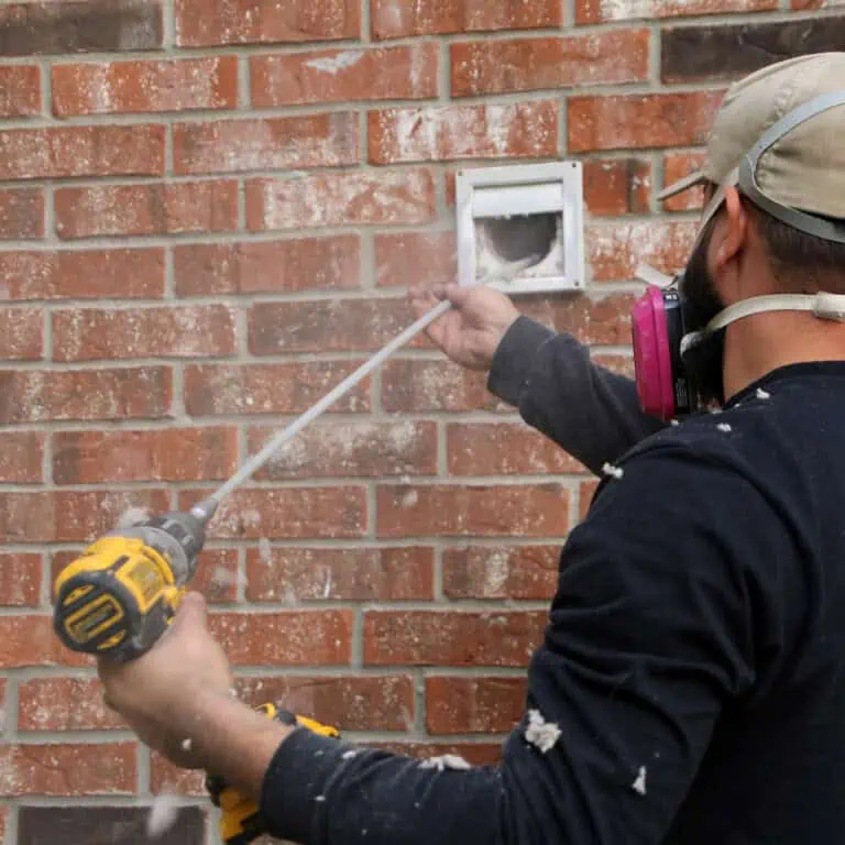 A person wearing a cap and respirator mask uses a power drill to cut into a brick wall, with dust flying and a square vent opening visible—typical of chimney repair work in Norfolk and Plymouth County, MA.