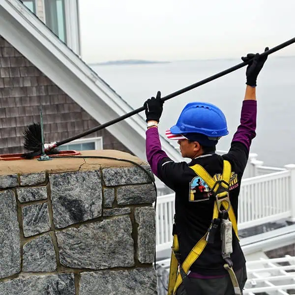 A worker in a blue hard hat and safety harness uses a long brush to clean a stone chimney on a rooftop near the water highlighting chimney repair expertise in Norfolk and Plymouth County, MA.