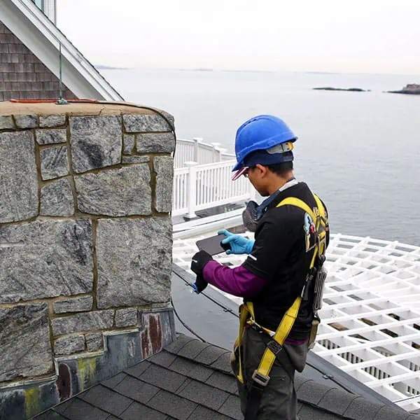 A construction worker in a blue hard hat and safety harness stands on a rooftop near the water in Norfolk and Plymouth County, MA, inspecting a stone chimney while holding a clipboard and wearing protective gloves.