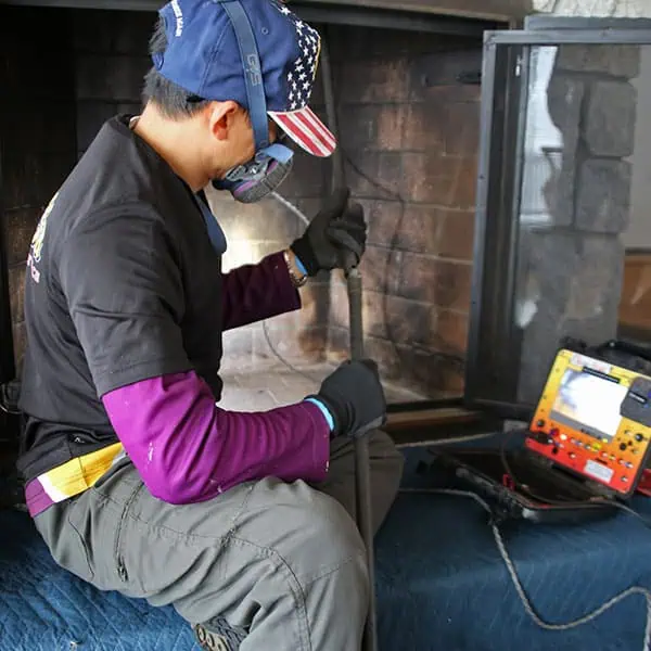 A person in protective gear and a cap with a U.S. flag design cleans a fireplace chimney using specialized tools, while operating a diagnostic machine beside the hearth—demonstrating expert chimney sweep services in Norfolk and Plymouth County, MA.