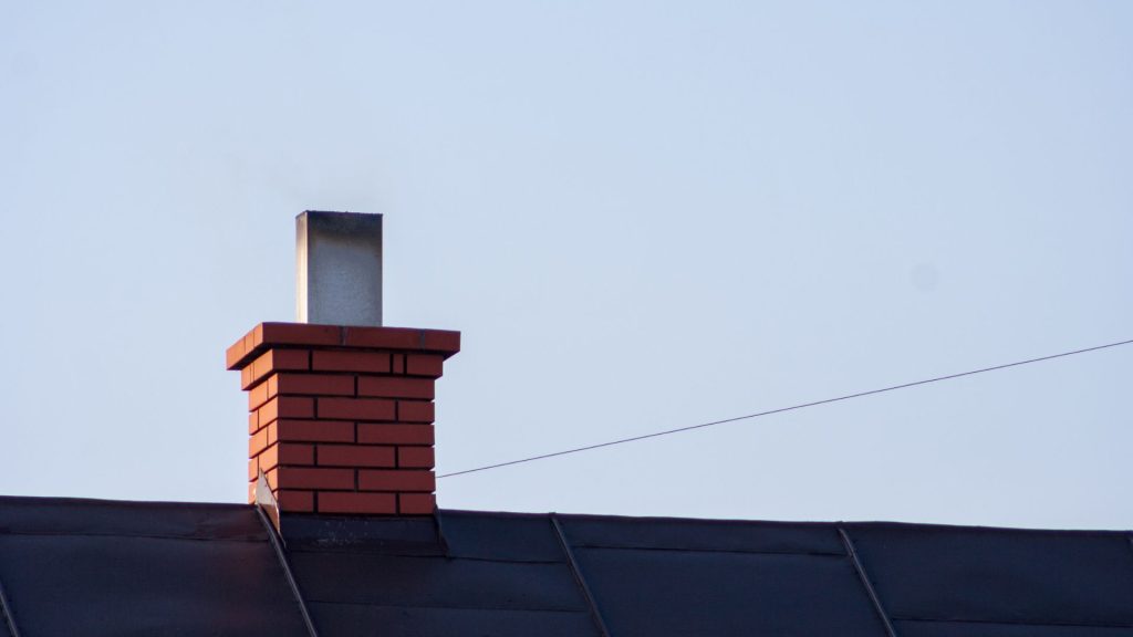 A brick chimney with light smoke rising sits on a dark rooftop against a clear blue MA sky, with a thin wire running across the background—ideal inspiration for chimney repair Norfolk and Plymouth County.