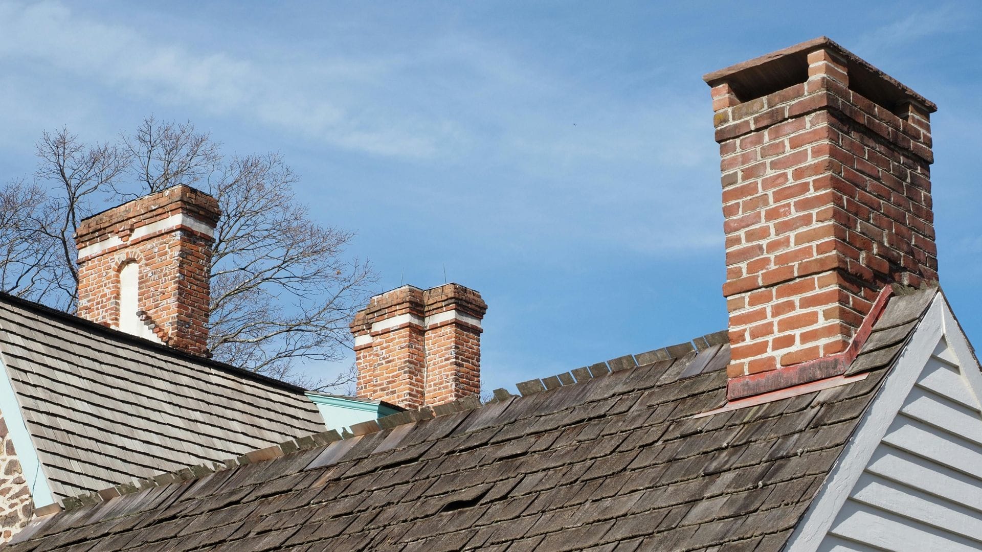 Three brick chimneys rise from the shingled rooftops of historic Massachusetts homes under a bright blue sky, with leafless tree branches in the background—evoking a typical scene for chimney sweep work in Norfolk and Plymouth County.