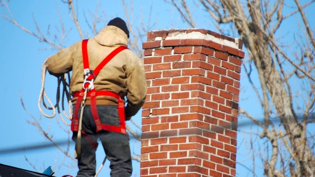 A worker wearing a safety harness and jacket stands on a roof next to a red brick chimney, holding a rope, during a chimney restoration in Norfolk and Plymouth County, MA. Leafless tree branches and a clear blue sky fill the background.