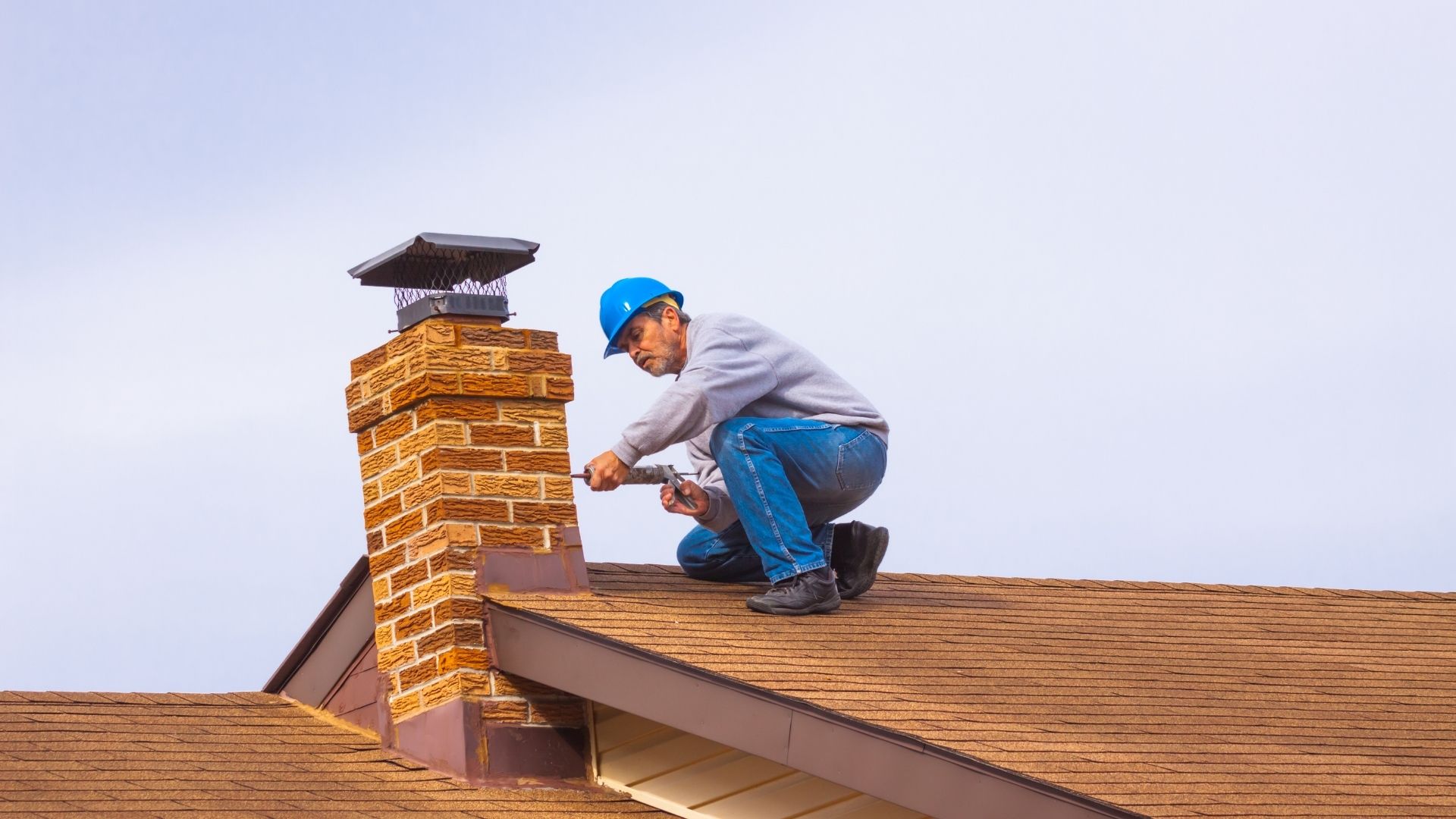 Worker in a blue hard hat and gray sweatshirt kneels on a sloped shingle roof, inspecting a brick chimney with tools under a clear blue sky in Norfolk and Plymouth County, MA.