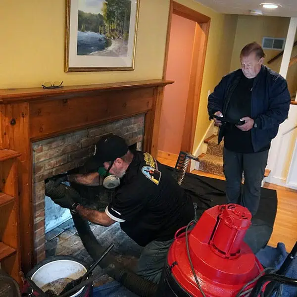 A man wearing a protective mask vacuums the inside of a brick fireplace using a hose, while another man stands nearby watching and holding a tablet, possibly assessing chimney repair services in Norfolk and Plymouth County, MA, inside a cozy living room.