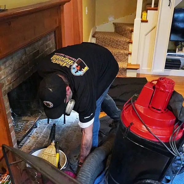 A worker wearing a cap and mask cleans a fireplace using a vacuum and cleaning tools, with protective materials arranged around the hearth—demonstrating expert chimney sweep Norfolk and Plymouth County, MA services.