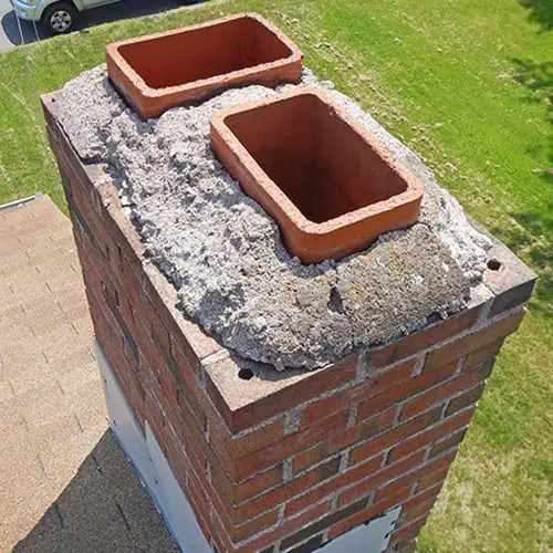 Close-up of a brick chimney with two rectangular clay flue liners in MA, surrounded by rough, uneven, and degraded concrete at the top, viewed from above on a sunny day. Grass and part of a rooftop are visible in the background.