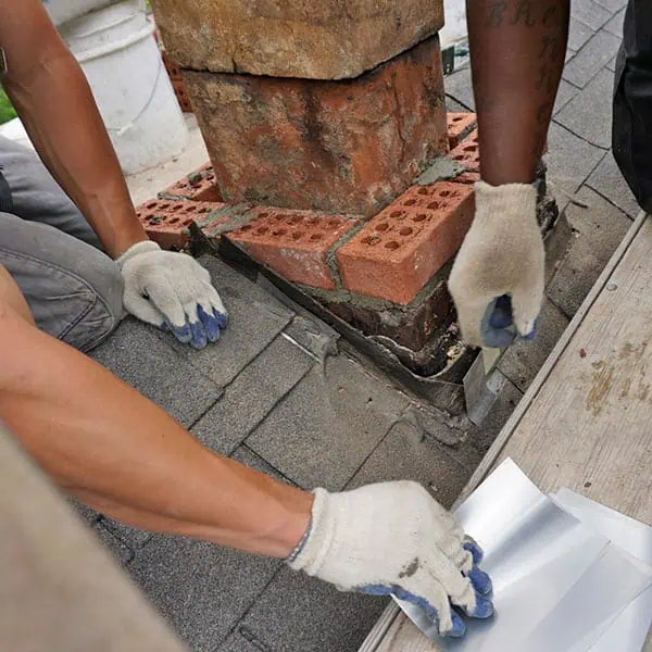Two people wearing work gloves install metal flashing around the base of a brick chimney on a shingled roof, securing the waterproof barrier to prevent leaks during chimney restoration in Norfolk and Plymouth County, MA.