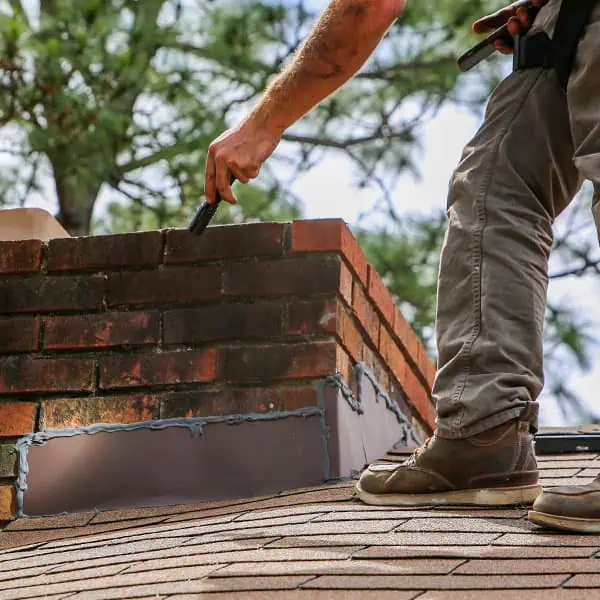 A person stands on a shingled roof using tools to repair the flashing and bricks around a brick chimney, performing chimney repair Norfolk and Plymouth County, MA, with trees visible in the background.
