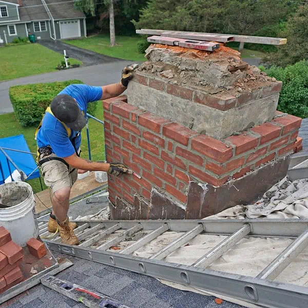 A worker in a blue shirt and safety gear repairs a brick chimney on a sloped roof in MA, surrounded by ladders and buckets—a scene typical of chimney repair in Norfolk and Plymouth County, with houses and greenery visible in the background.