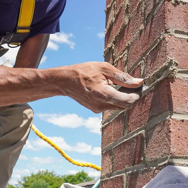A person uses a trowel to repair mortar between bricks on an outdoor brick wall, showcasing expert chimney restoration in Norfolk and Plymouth County, with a bright blue sky in the background.