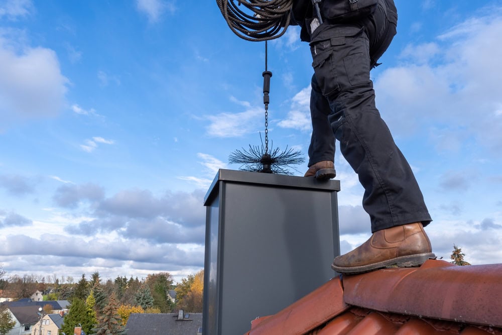 A person in black pants and brown boots stands on a sloped red roof, using a chimney cleaning brush attached to a rod to sweep a rectangular chimney under partly cloudy skies—demonstrating chimney sweep work in Norfolk and Plymouth County, MA.