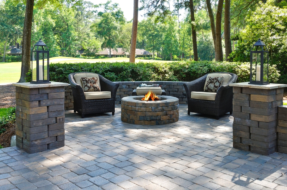 A patio in MA with two wicker armchairs and cushions faces a stone fire pit, surrounded by brick pillars with lanterns—perfect for relaxing after expert chimney repair Norfolk and Plymouth County services in this lush, green, wooded area.