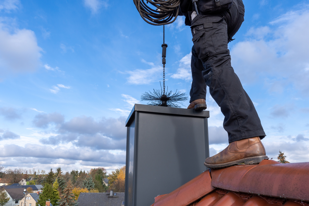 A person stands on a rooftop, using a long cable brush to clean a chimney, with clouds and trees in the background—chimney sweep Norfolk and Plymouth County, MA.