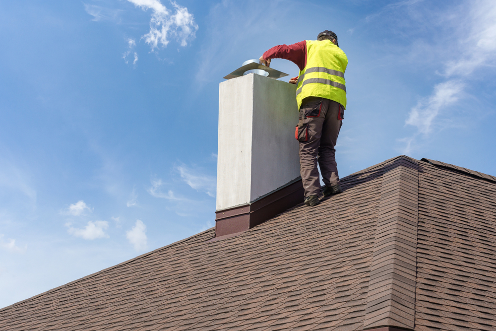 A worker in a yellow safety vest stands on a shingled roof, inspecting or repairing a chimney under a blue sky in Norfolk and Plymouth County, MA.