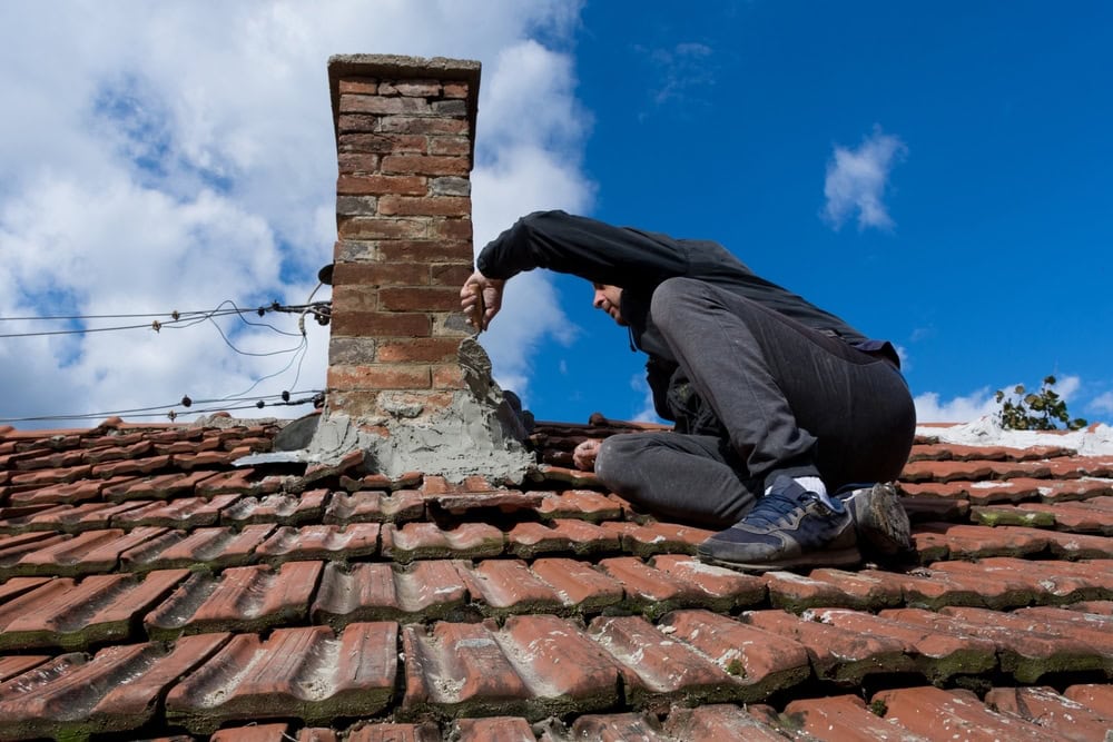 A person dressed in dark work clothing kneels carefully on a sloped, red-tiled roof while applying fresh mortar to the base of a weathered brick chimney. The individual’s posture and tools suggest precise masonry work and skilled craftsmanship. The background reveals a soft blue sky with scattered white clouds, indicating a clear and calm day—ideal conditions for rooftop chimney repair. This scene reflects professional chimney maintenance commonly required for homes in Norfolk and Plymouth County, Massachusetts.