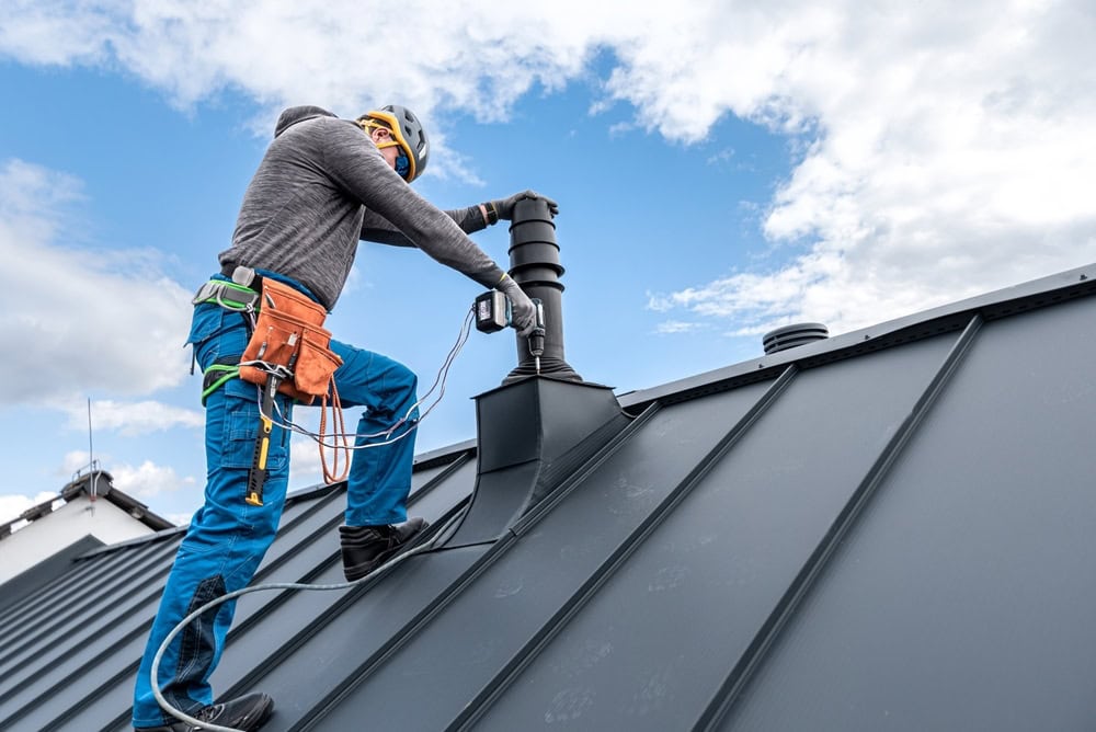 A worker in safety gear and a tool belt uses a power drill on a sloped metal roof under a partly cloudy sky, assisting with chimney restoration Norfolk and Plymouth County, MA.