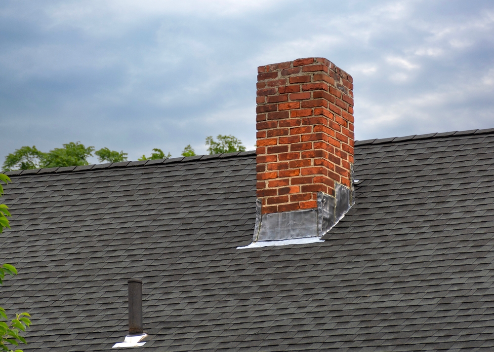 A red brick chimney with metal flashing extends from a gray shingled roof on a Massachusetts home, surrounded by trees and under a cloudy sky—an example of expert chimney restoration in Norfolk and Plymouth County, MA.