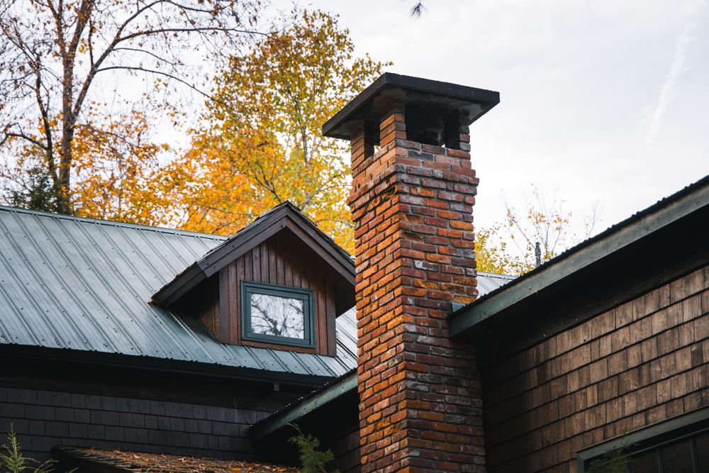 A brick chimney rises from the roof of a wooden house with a metal roof, surrounded by trees with autumn-colored leaves under a cloudy sky—an ideal setting for expert chimney repair Norfolk and Plymouth County, MA.