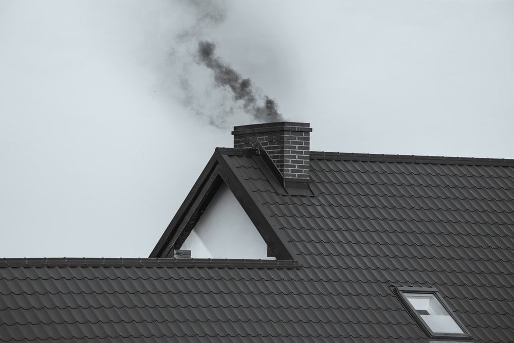 Dark smoke rises from a red-brick chimney on a tiled house roof against a gray sky, suggesting the fireplace or stove is in use. Homeowners seeking chimney repair in Norfolk and Plymouth County, MA will recognize this classic scene. A skylight window is visible nearby.