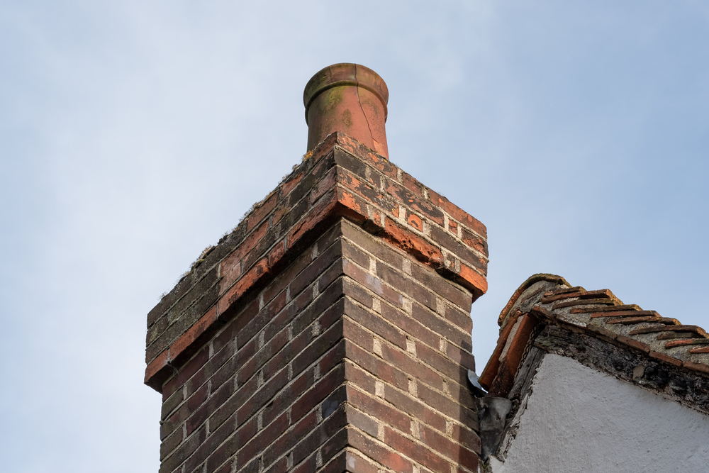Close-up of a brick chimney featuring a weathered red clay chimney pot on a tiled roof, with blue sky in the background—serviced by a professional chimney sweep in Norfolk and Plymouth County, MA.
