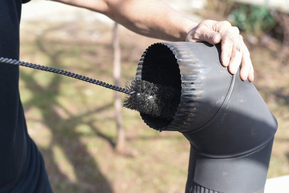 A chimney sweep wearing black pants and brown boots stands securely on a sloped red metal roof, using a long rod and brush to clean the inside of a rectangular chimney under a partly cloudy sky.