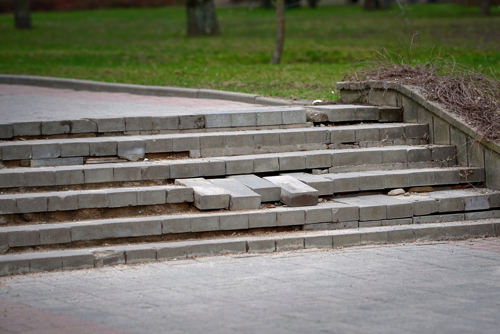A set of outdoor concrete steps with several broken and uneven bricks, creating a hazardous surface—much like damaged chimneys that require chimney restoration Norfolk and Plymouth County. Grass and trees are visible in the background.