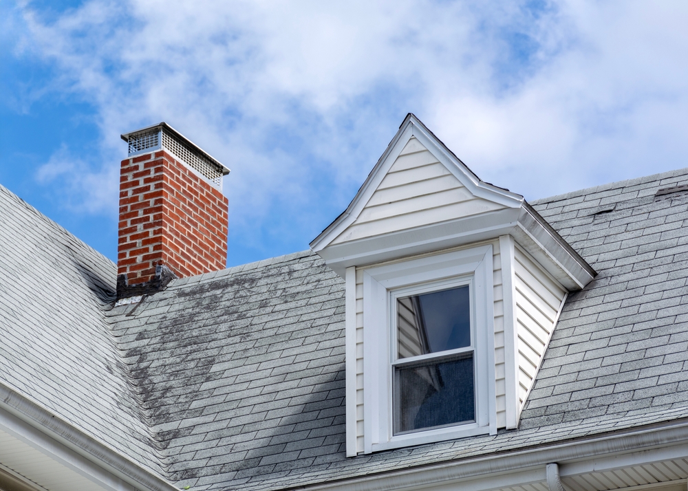 A red brick chimney rises from a gray-shingled roof with a white dormer window under a blue sky with scattered clouds representing a classic Massachusetts home ideal for chimney restoration in Norfolk and Plymouth County.