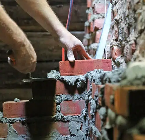Close-up of hands applying mortar and laying red bricks on a partially completed wall, with construction tools nearby representing chimney repair in Norfolk and Plymouth County, MA.
