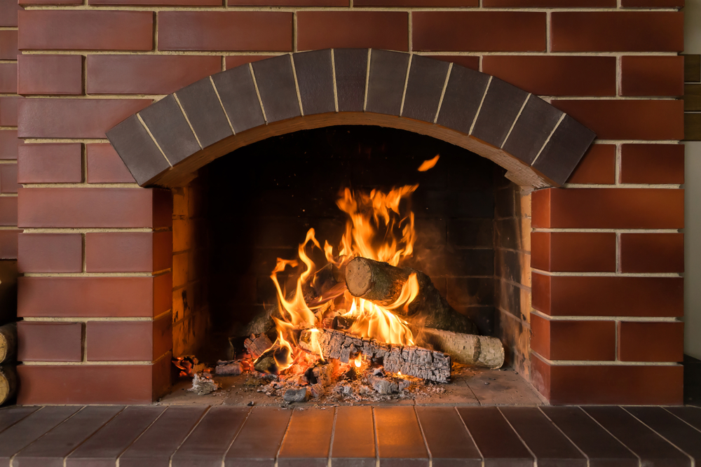 A fire burns in a brick fireplace, warming the MA home with orange flames rising around stacked logs and embers glowing on the hearth. The reddish-brown bricks with light grout lines add cozy charm to this MA setting.