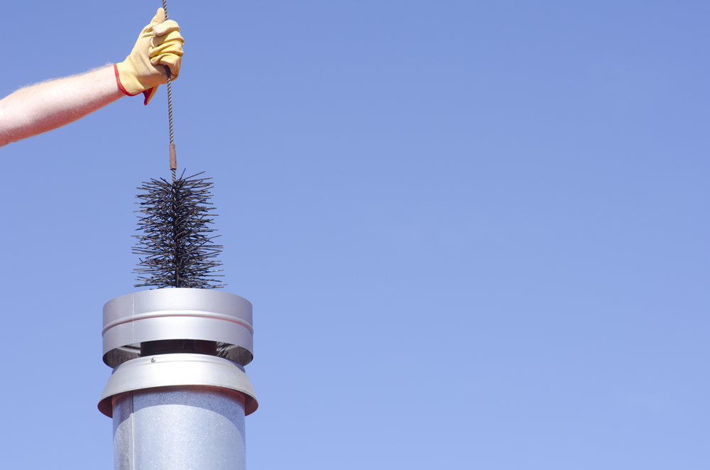 A person wearing a yellow glove is holding a chimney brush above a metal chimney pipe against a clear blue sky, preparing for chimney repair Norfolk and Plymouth County, MA.