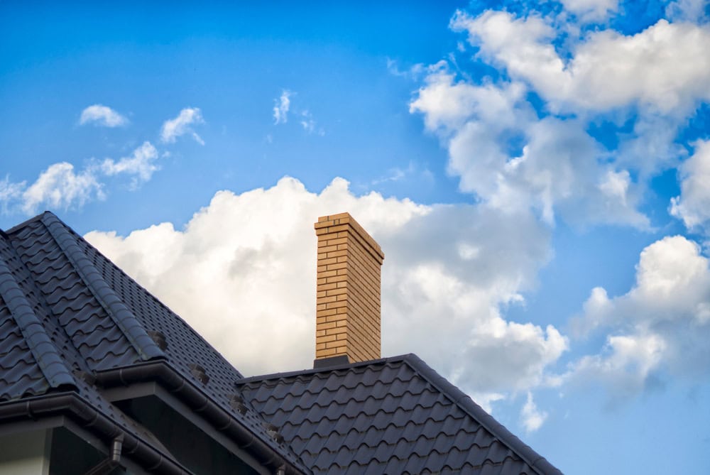 A brick chimney rises above a dark gray tiled roof in MA, set against a bright blue sky with scattered white clouds—perfect for a professional chimney sweep in Norfolk and Plymouth County, MA.