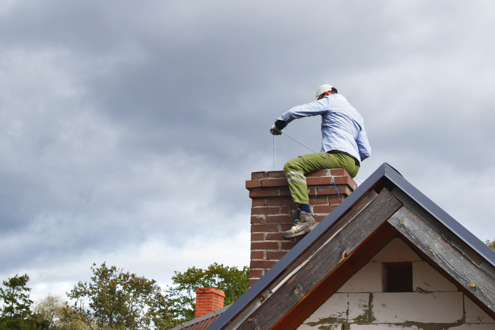 A person in a white cap and green pants is cleaning or inspecting a brick chimney on a roof under a cloudy sky, possibly performing chimney repair Norfolk and Plymouth County. Trees are visible in the background.