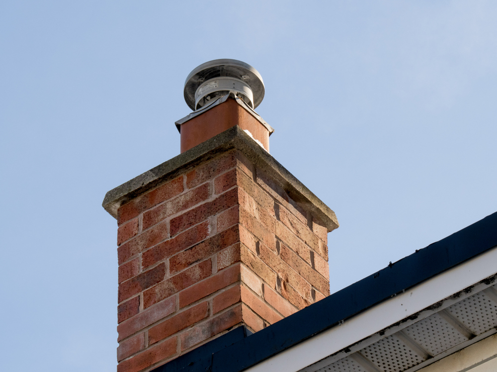 A red brick chimney with a metal cap on top, restored by experts in chimney restoration Norfolk and Plymouth County, rises above a rooftop against the clear blue MA sky.